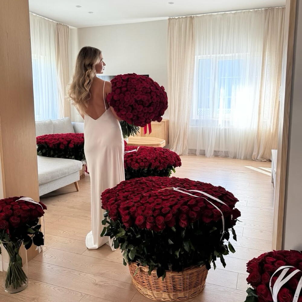 Woman in a white dress holding a large bouquet of red roses in a room with similar arrangements.